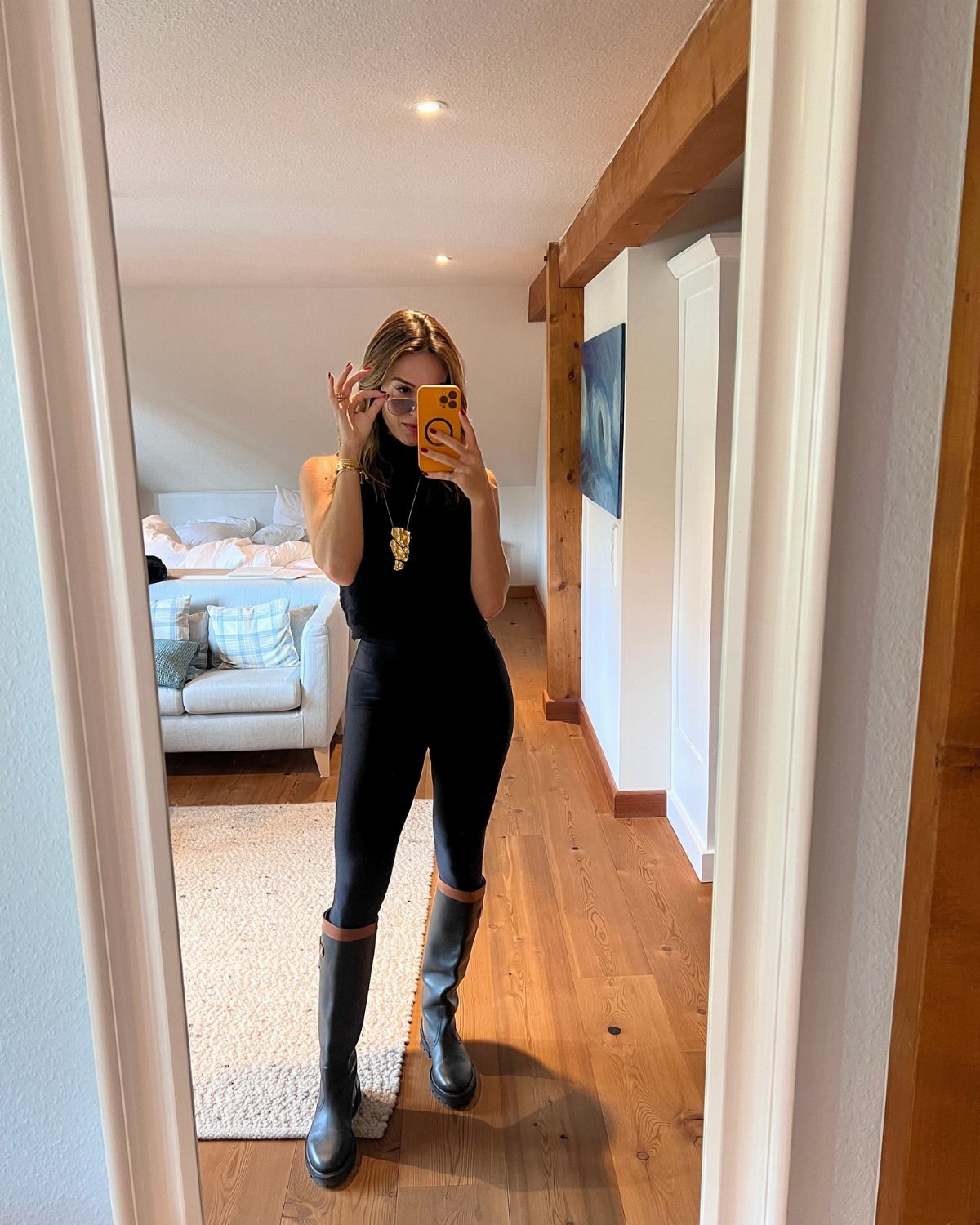 Woman in black leggings, sleeveless top, and tall black boots taking a mirror selfie.