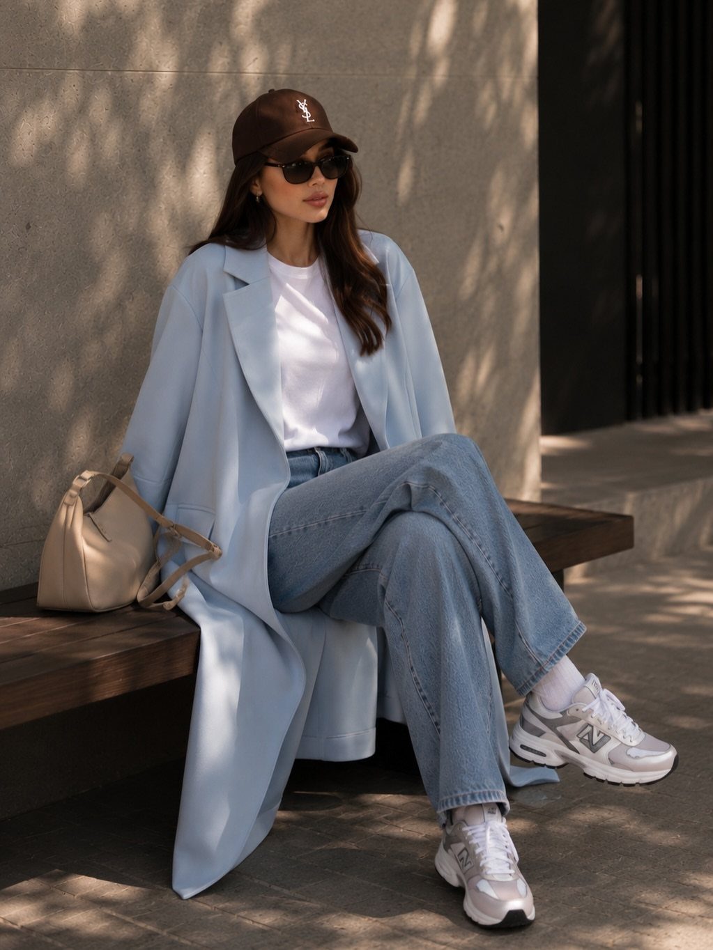 Woman in a light blue overcoat, white t-shirt, and blue jeans sitting on a bench.