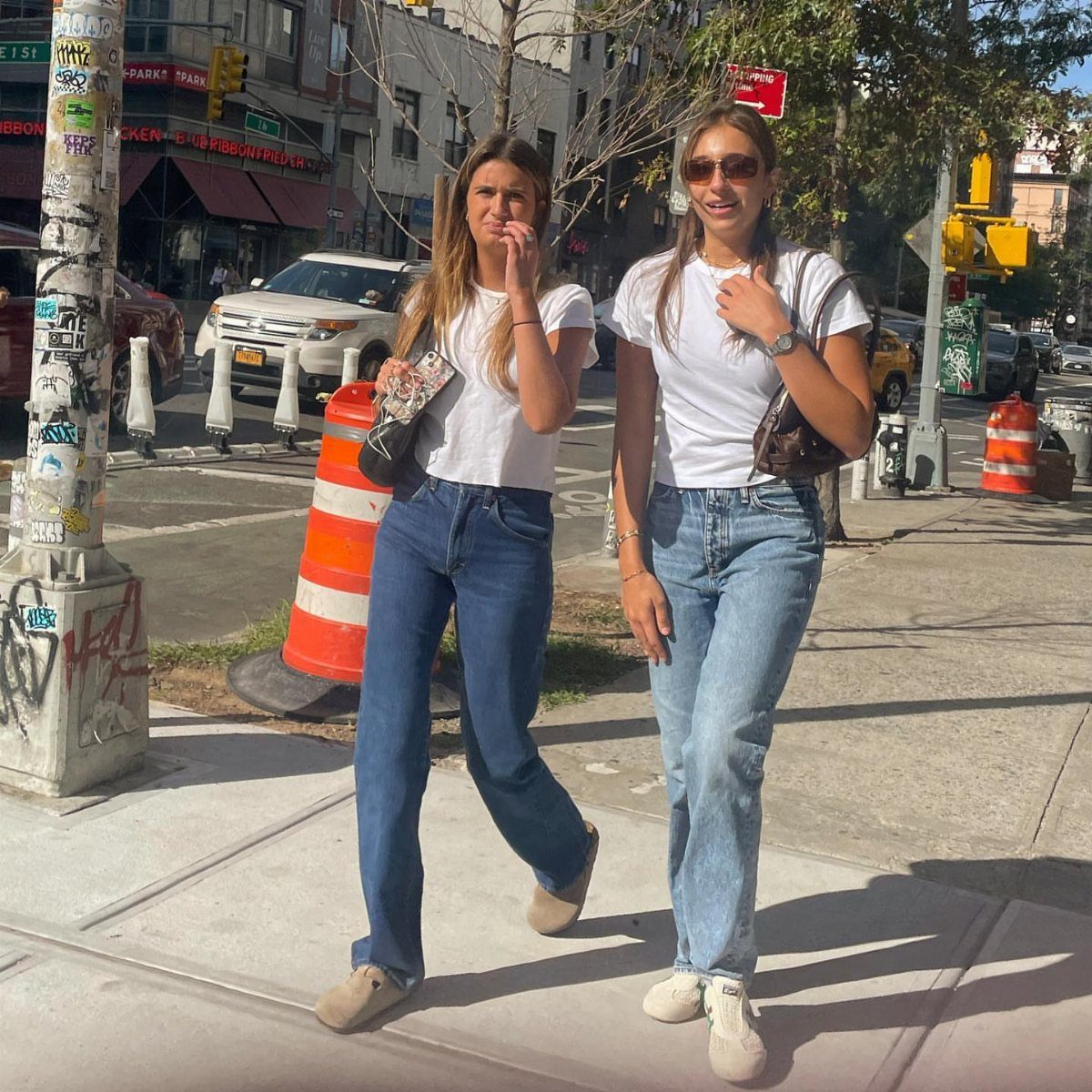 Two women walking on a city sidewalk wearing white t-shirts and blue jeans.