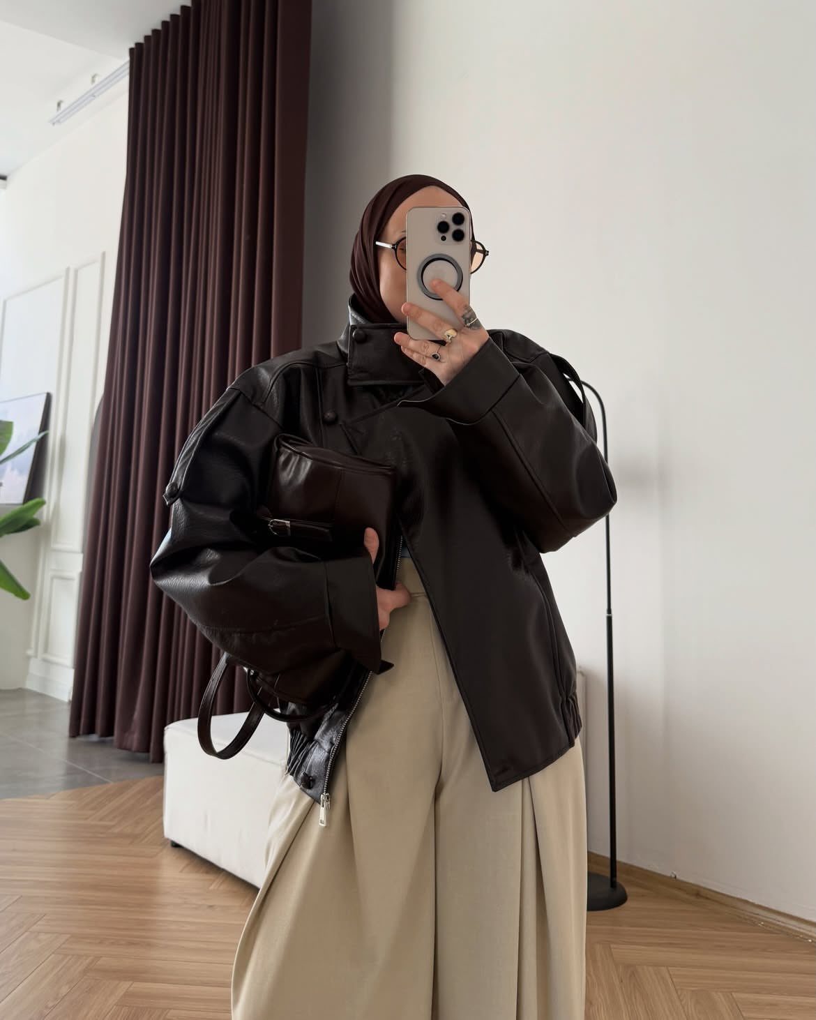Person in an oversized brown leather jacket and beige wide-leg trousers taking a mirror selfie.