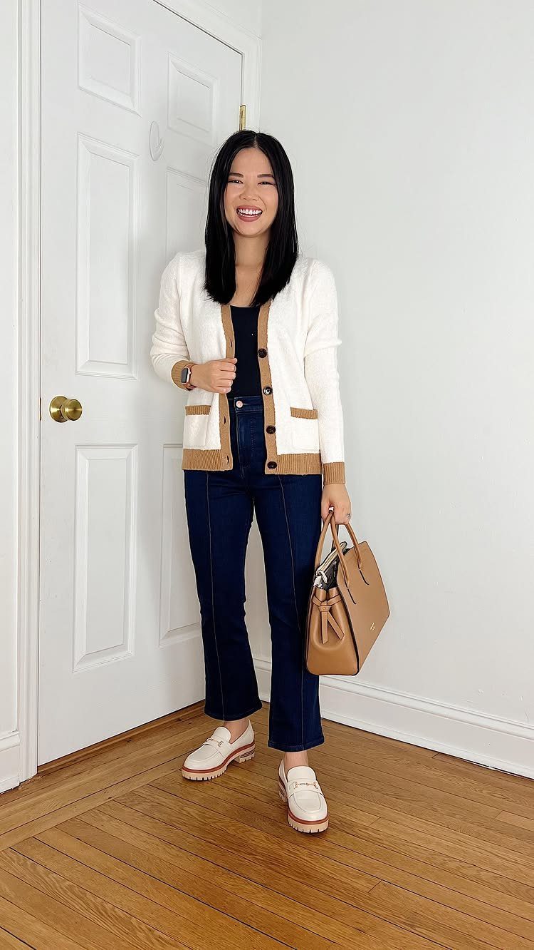 A smiling woman posing in a white cardigan, dark flared jeans, and white loafers holding a tan handbag.