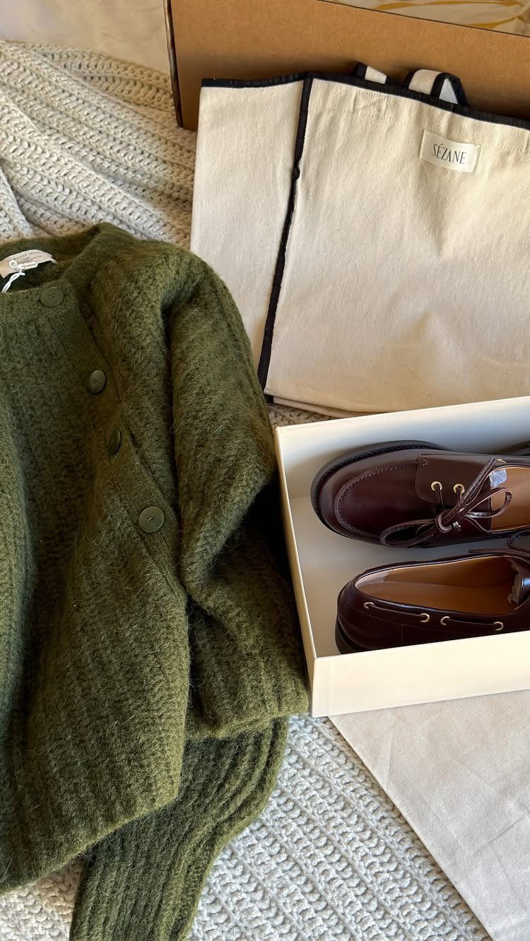 Olive green knit cardigan, brown leather loafers, and a cream canvas tote bag on white bedding.