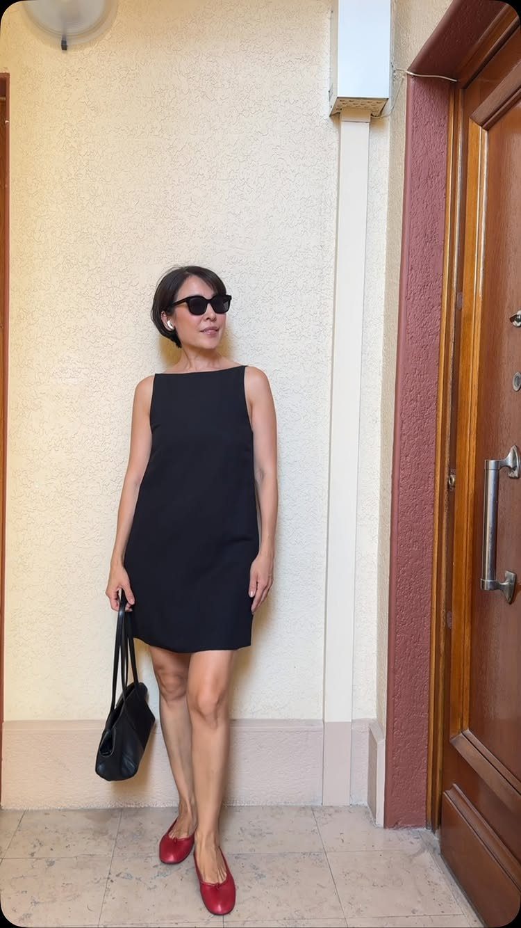 A woman in a sleeveless black dress, sunglasses, and red flats standing by a door.