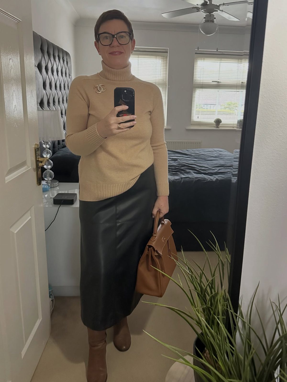 A woman in a tan turtleneck and black leather skirt posing for a mirror selfie.