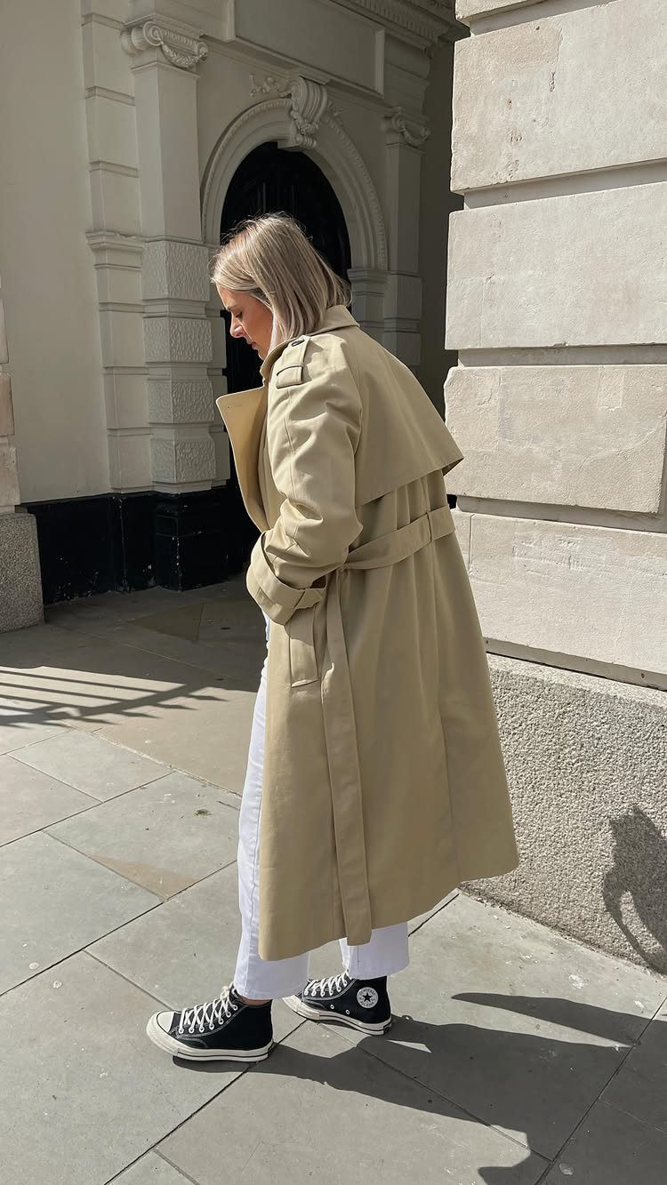 A woman wearing a long beige trench coat and white trousers standing by a stone building.