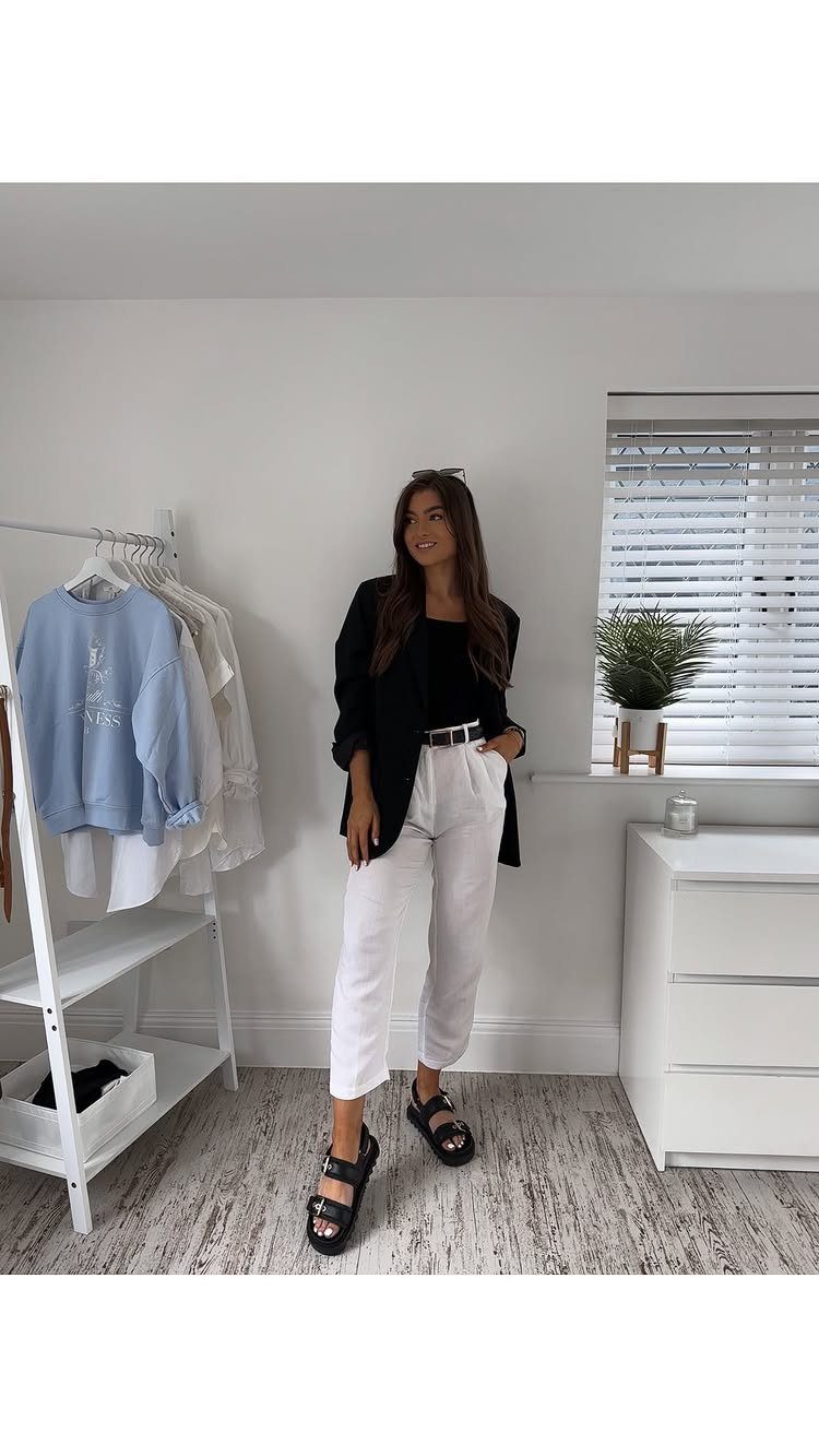 Woman wearing a black blazer, black top, and white trousers standing in a bright room.