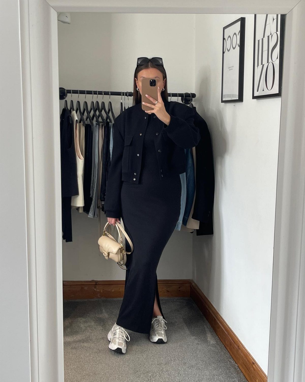Woman in a black maxi dress and jacket posing for a mirror selfie in a closet.