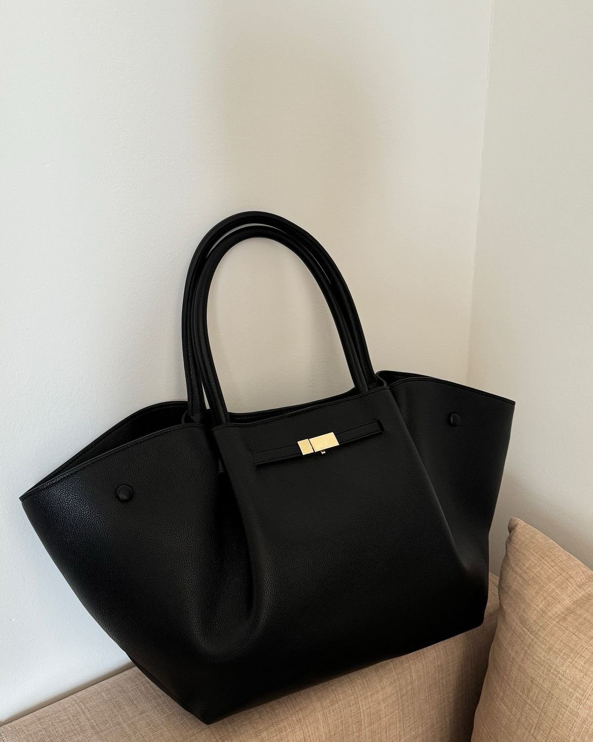 A black leather tote bag with gold hardware resting on a beige cushion.