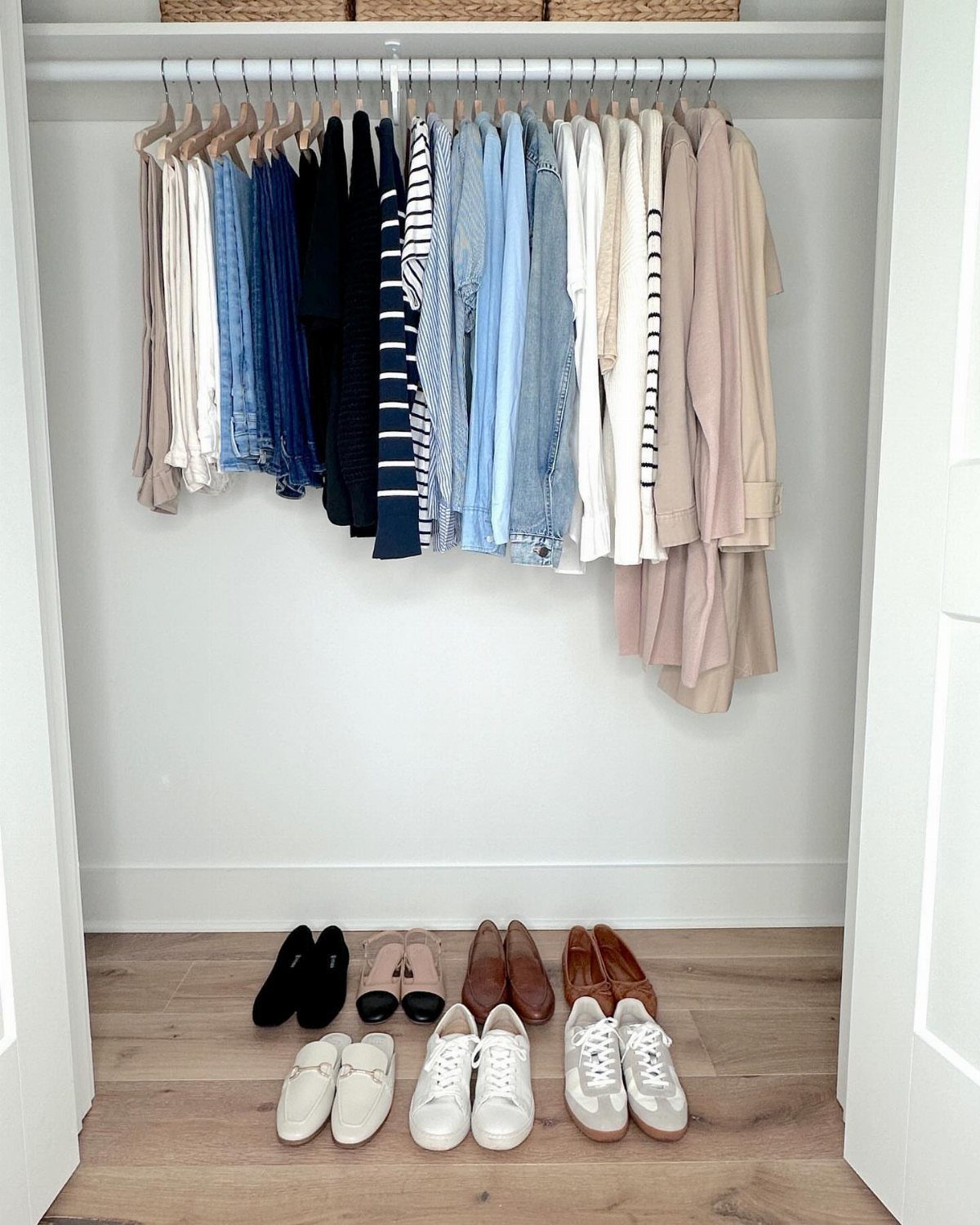 Clothing in neutral and blue tones hanging in a closet above various pairs of shoes.