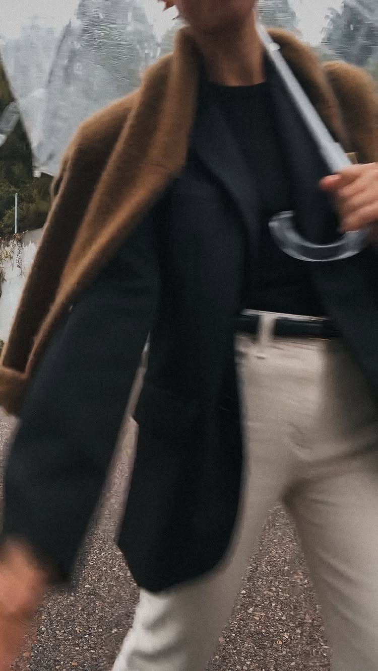 A woman walking outdoors wearing a black blazer, brown coat, and light trousers while holding an umbrella.