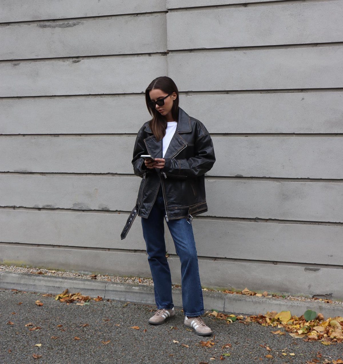 A woman wearing a black leather jacket, white shirt, and blue jeans standing against a wall.