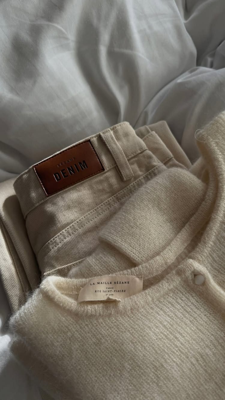 Cream knit sweater and folded denim trousers resting on white wrinkled bedding.