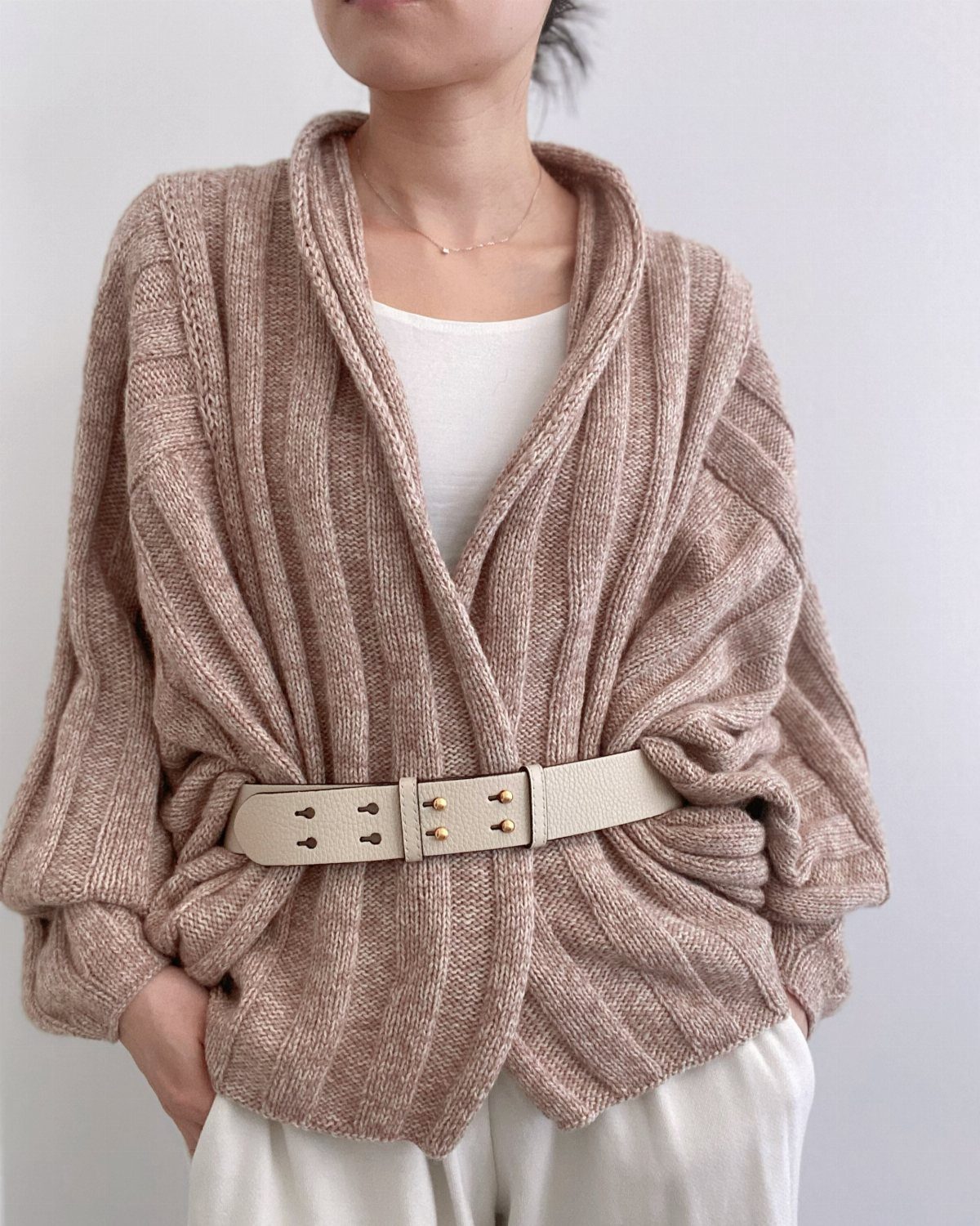 A person wearing a textured beige chunky knit cardigan cinched with a wide cream belt.