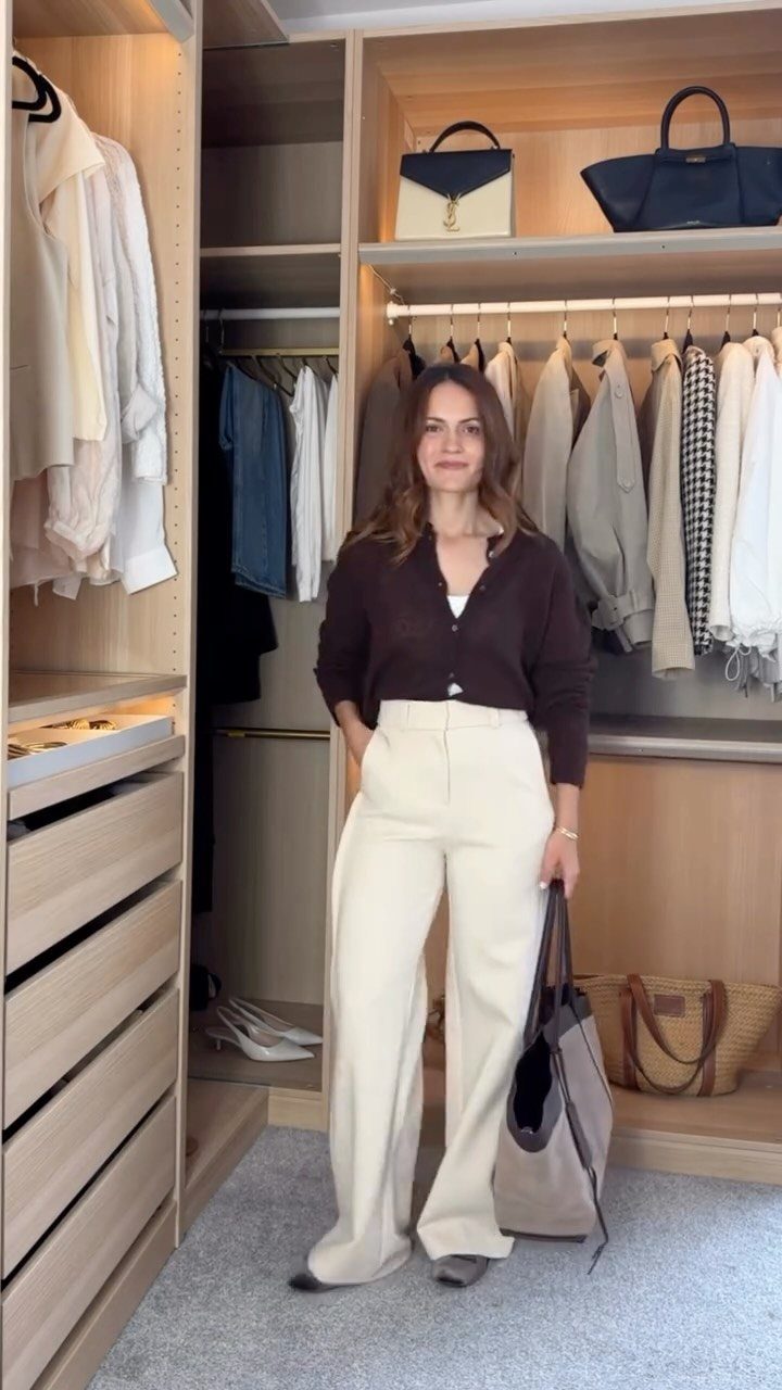 A woman posing in a walk-in closet wearing a brown ribbed knit cardigan and cream trousers.