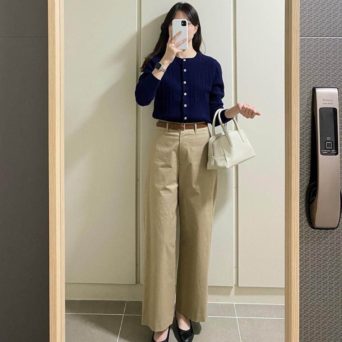 Woman in navy cardigan and tan trousers posing for a mirror selfie.