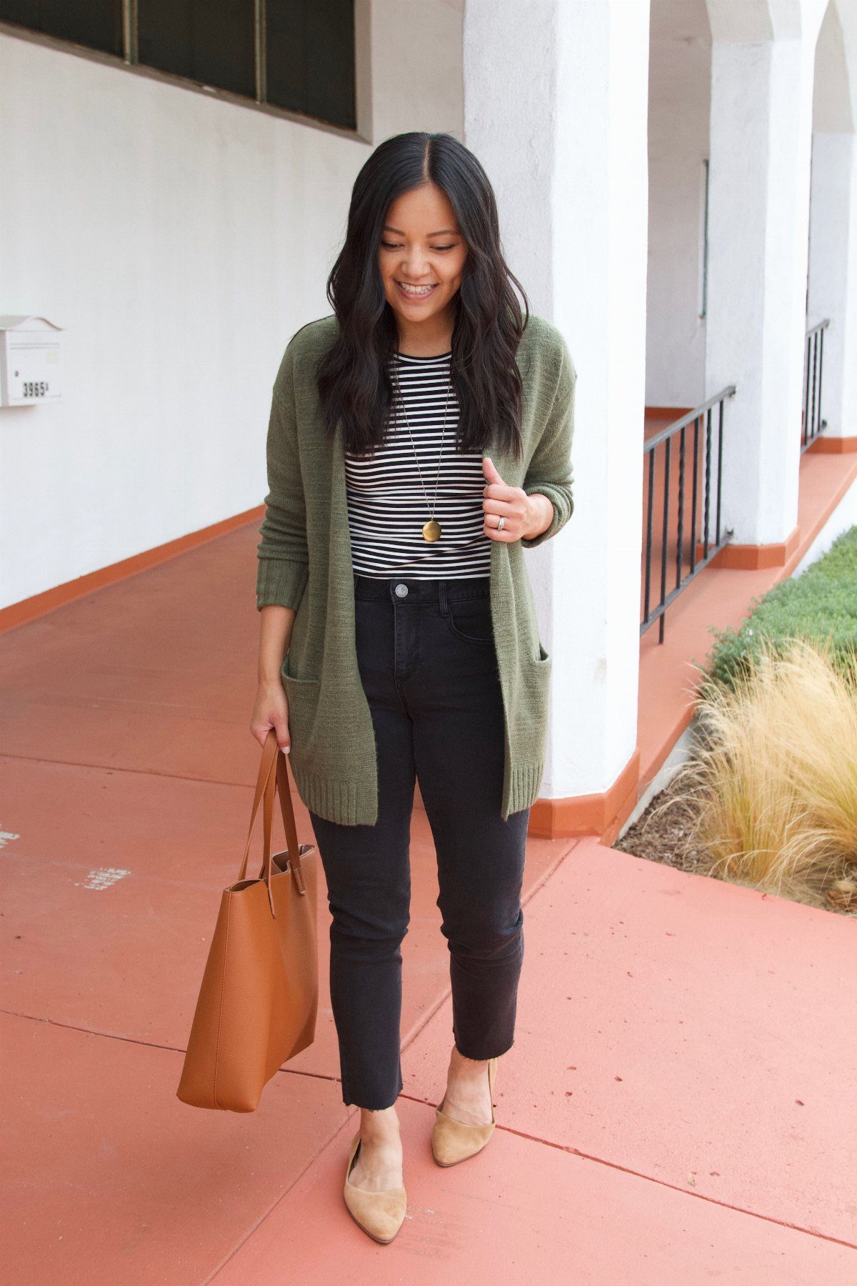 A woman wearing an olive green knit cardigan, striped shirt, black pants, and tan flats walking outdoors.