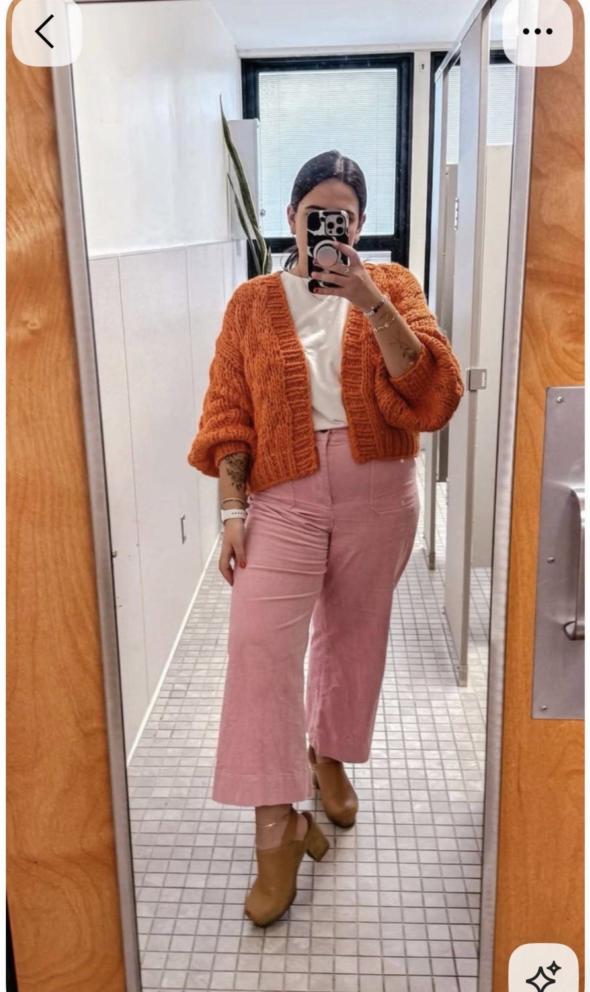 Woman in an orange chunky knit cardigan, white top, and pink trousers posing for a mirror selfie.