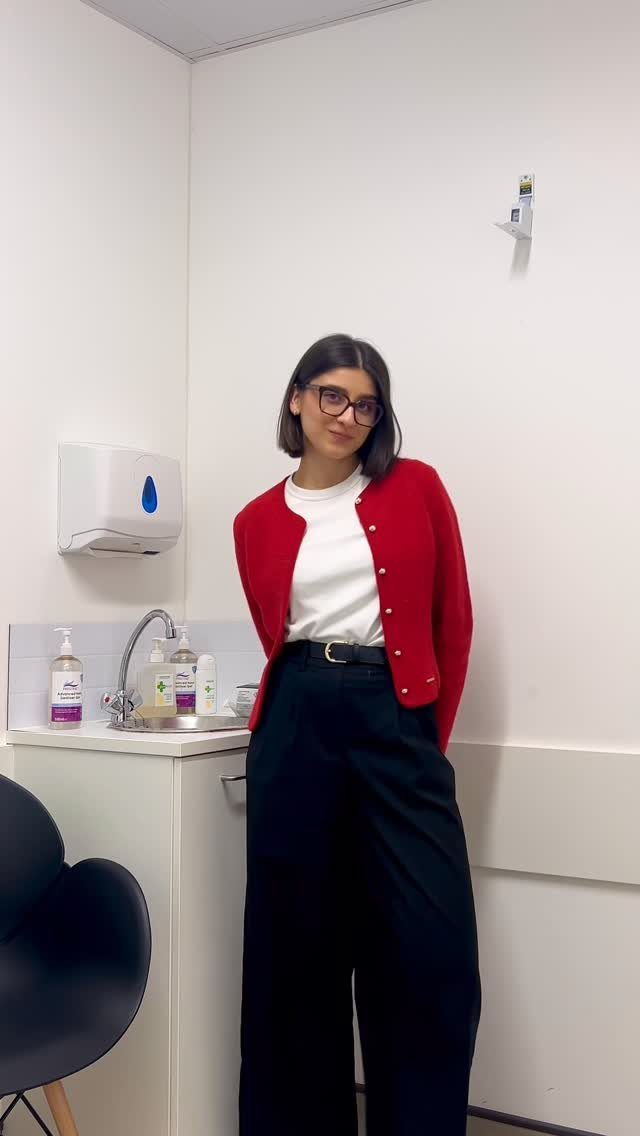 Woman wearing a red button-up cardigan, white shirt, and black trousers standing in a room.
