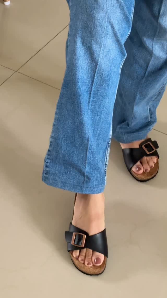 Person wearing blue wide-leg jeans and black buckled slide sandals on a tiled floor.