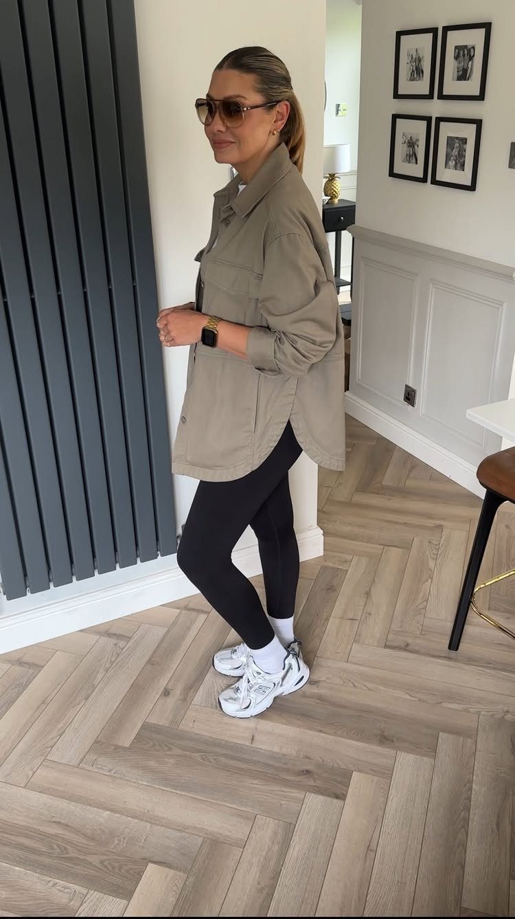 A woman wearing an oversized tan shirt, black leggings, and white sneakers standing indoors.