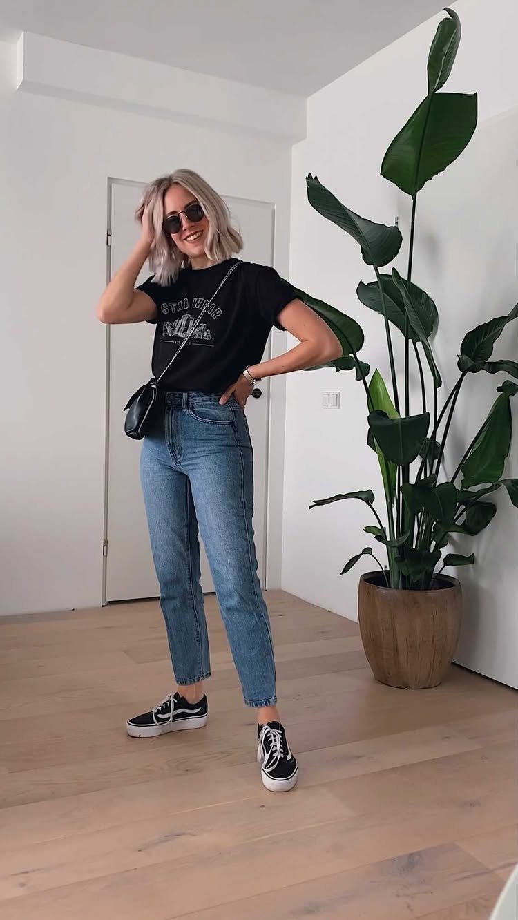 A woman posing in a black graphic t-shirt and blue denim jeans next to a plant.