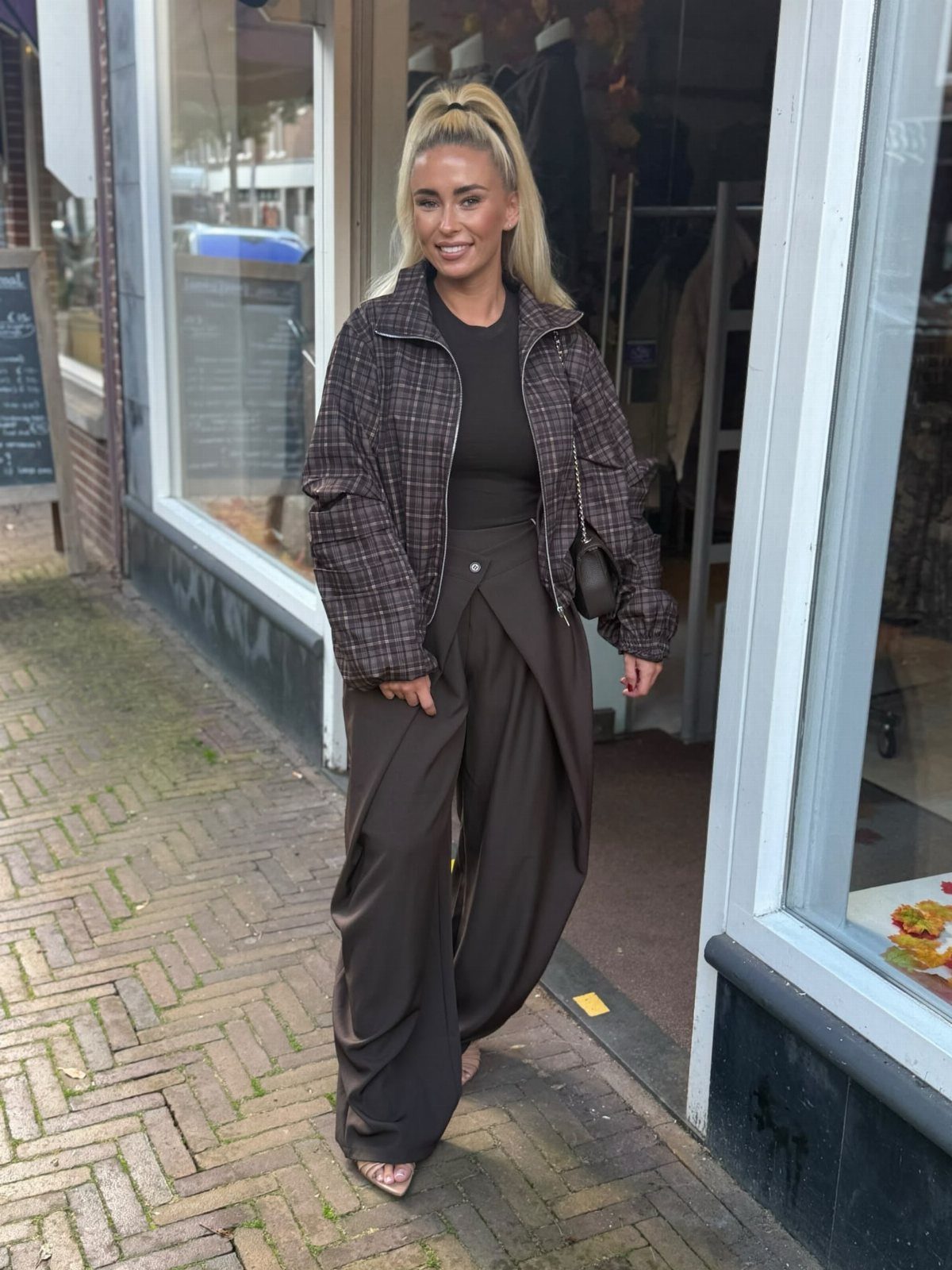 A blonde woman posing in a dark plaid jacket and brown wide-leg pants on a sidewalk.
