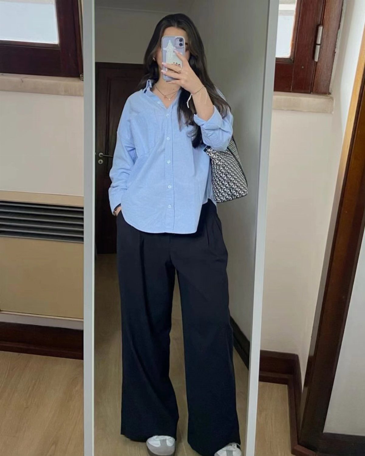 Woman in a light blue button-down shirt and black wide-leg trousers taking a mirror selfie.