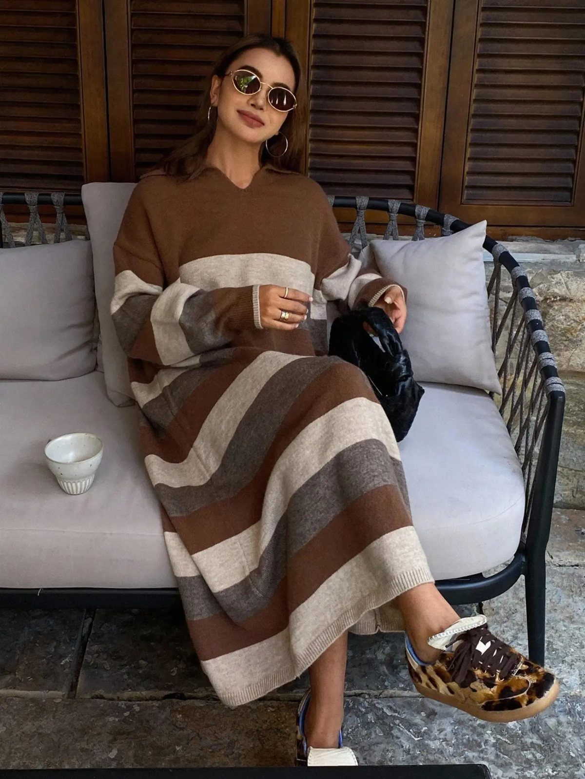 Woman sitting on an outdoor sofa wearing a long striped knit sweater dress and sunglasses.