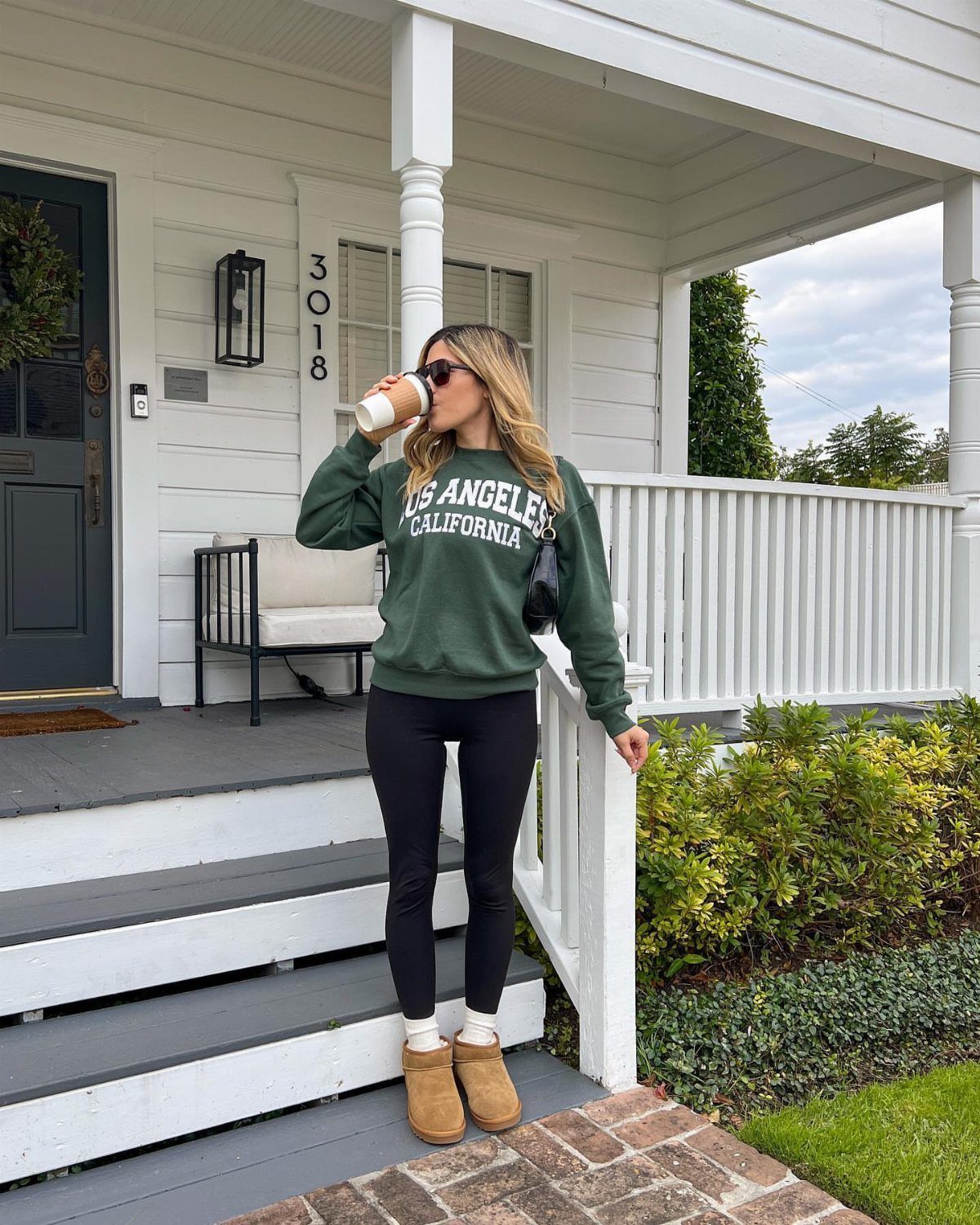 Woman in a green sweatshirt, black leggings, and tan boots standing on a white porch.