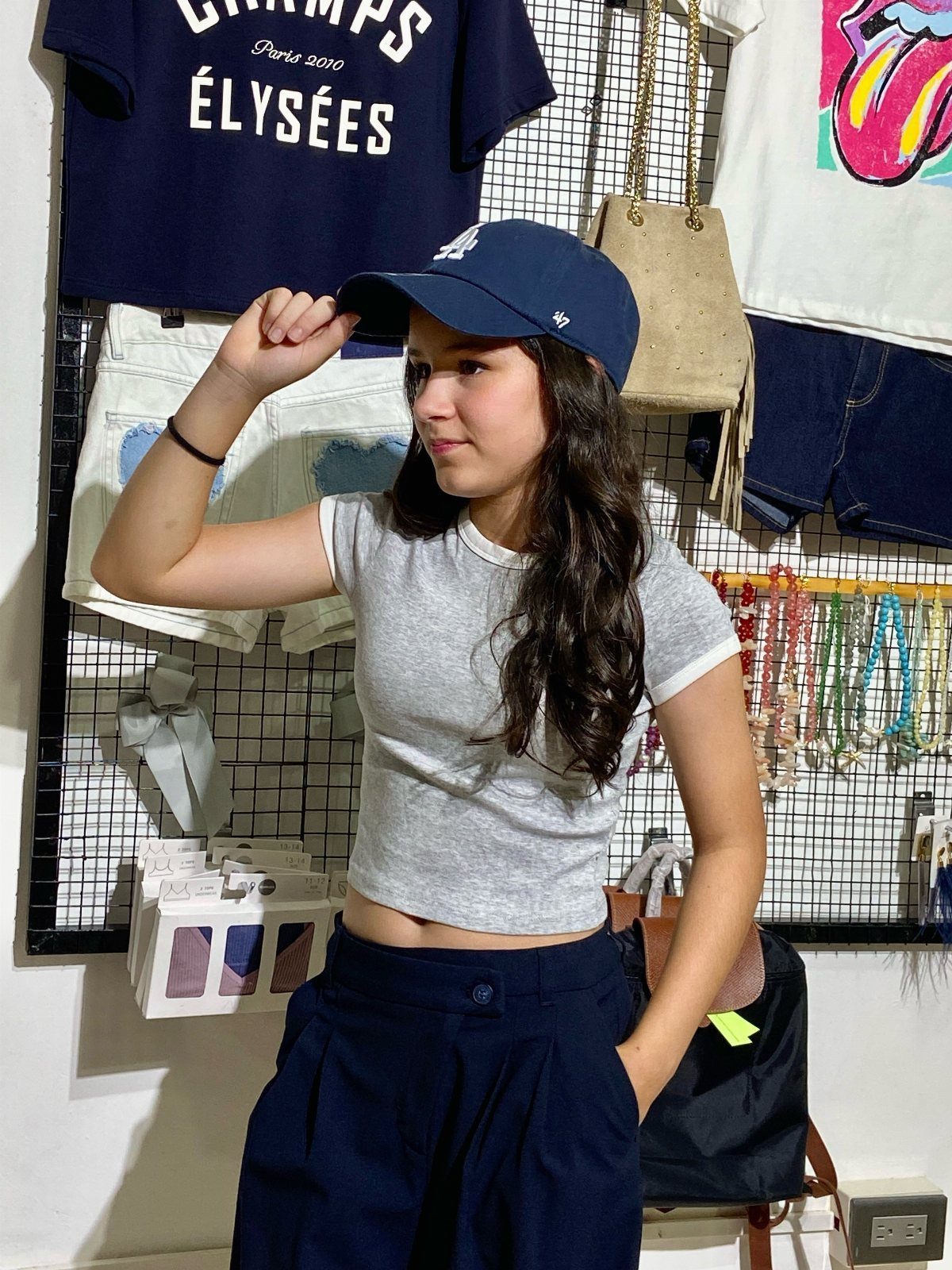 A teenage girl in a grey crop top and navy cap posing in a clothing store.