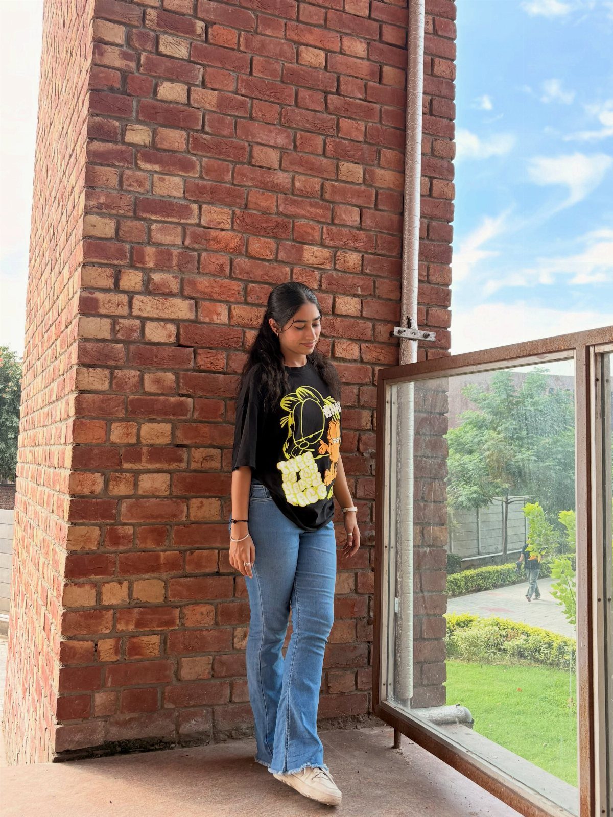 A young woman wearing a black graphic t-shirt and blue flared jeans standing by a brick wall.