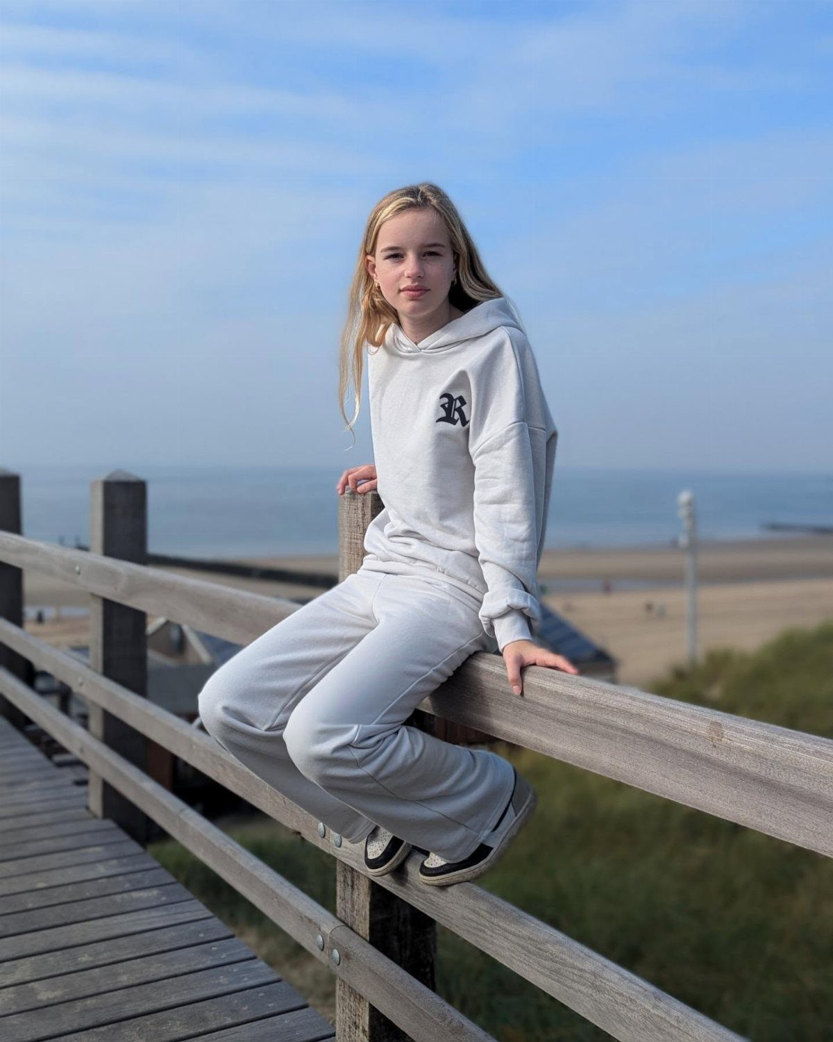 A blonde girl sitting on a wooden fence overlooking a beach in a white tracksuit.