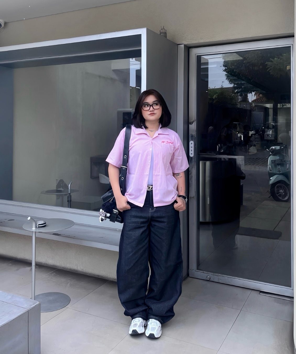 A young woman wearing an oversized pink shirt and wide-leg dark jeans standing outdoors.