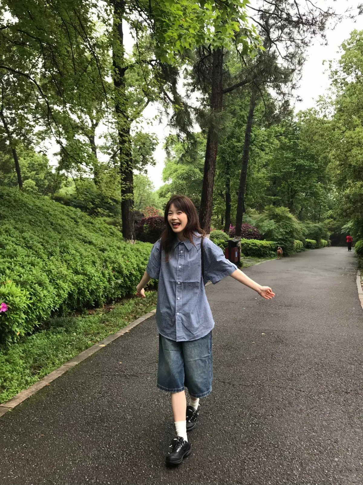 A young woman walking through a green park wearing an oversized blue checkered shirt and denim shorts.