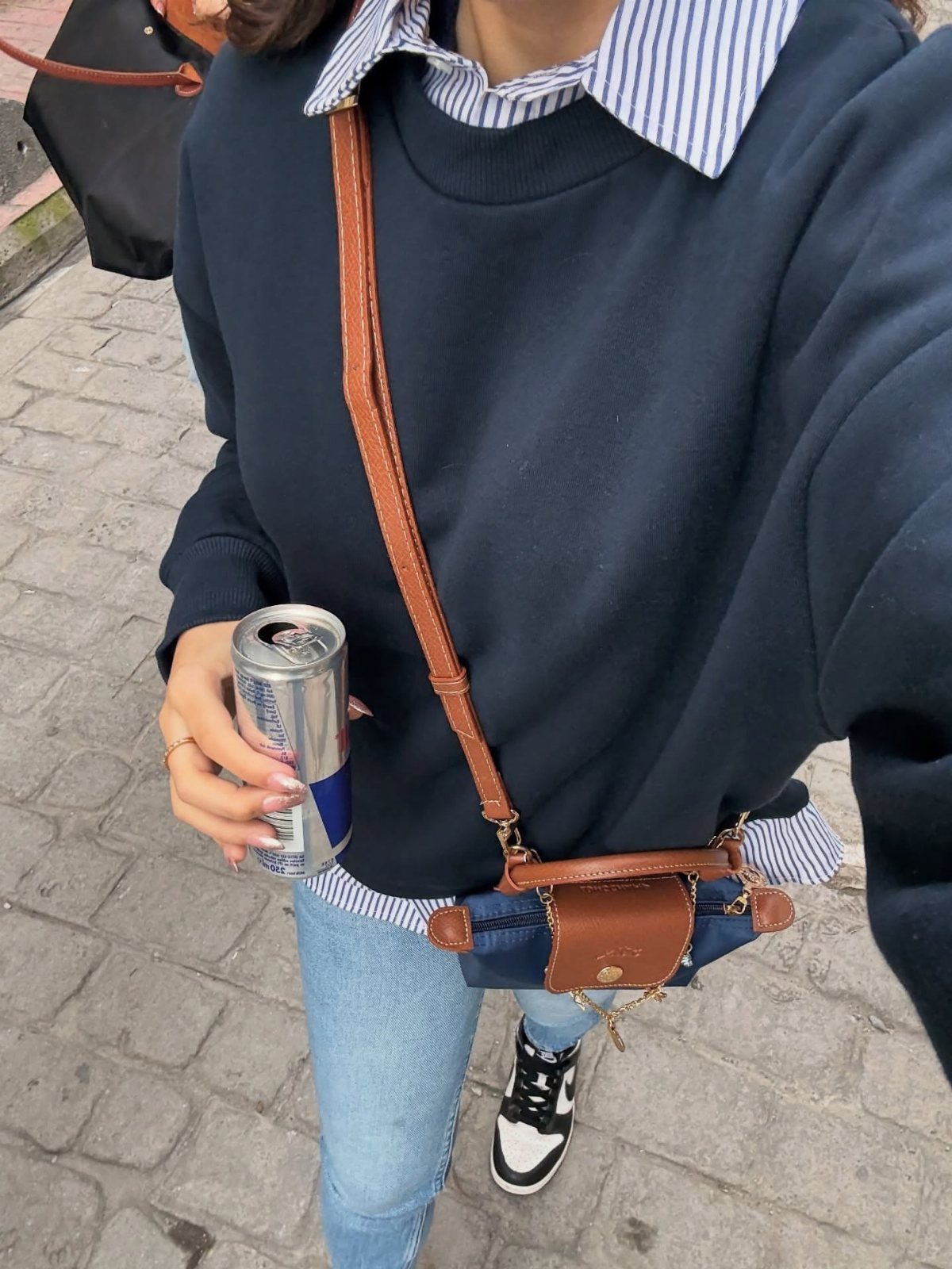 Person wearing a navy sweatshirt, striped shirt, and light blue denim jeans while holding a can.