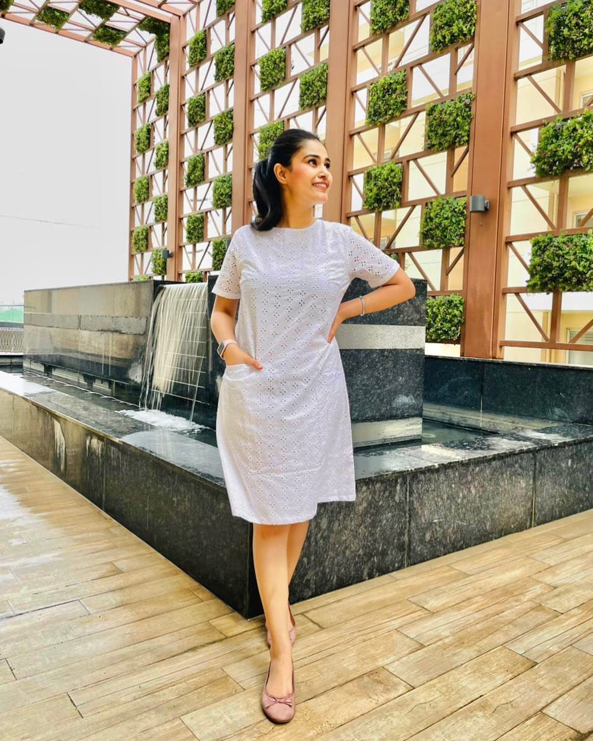 A young woman posing in a short-sleeved white woven dress near a stone fountain.