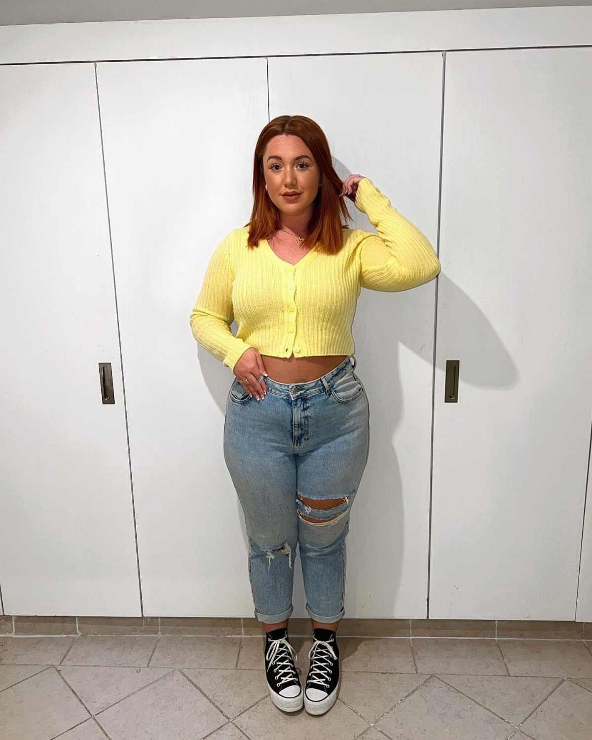 A young woman wearing a yellow cardigan, ripped jeans, and black sneakers posing indoors.