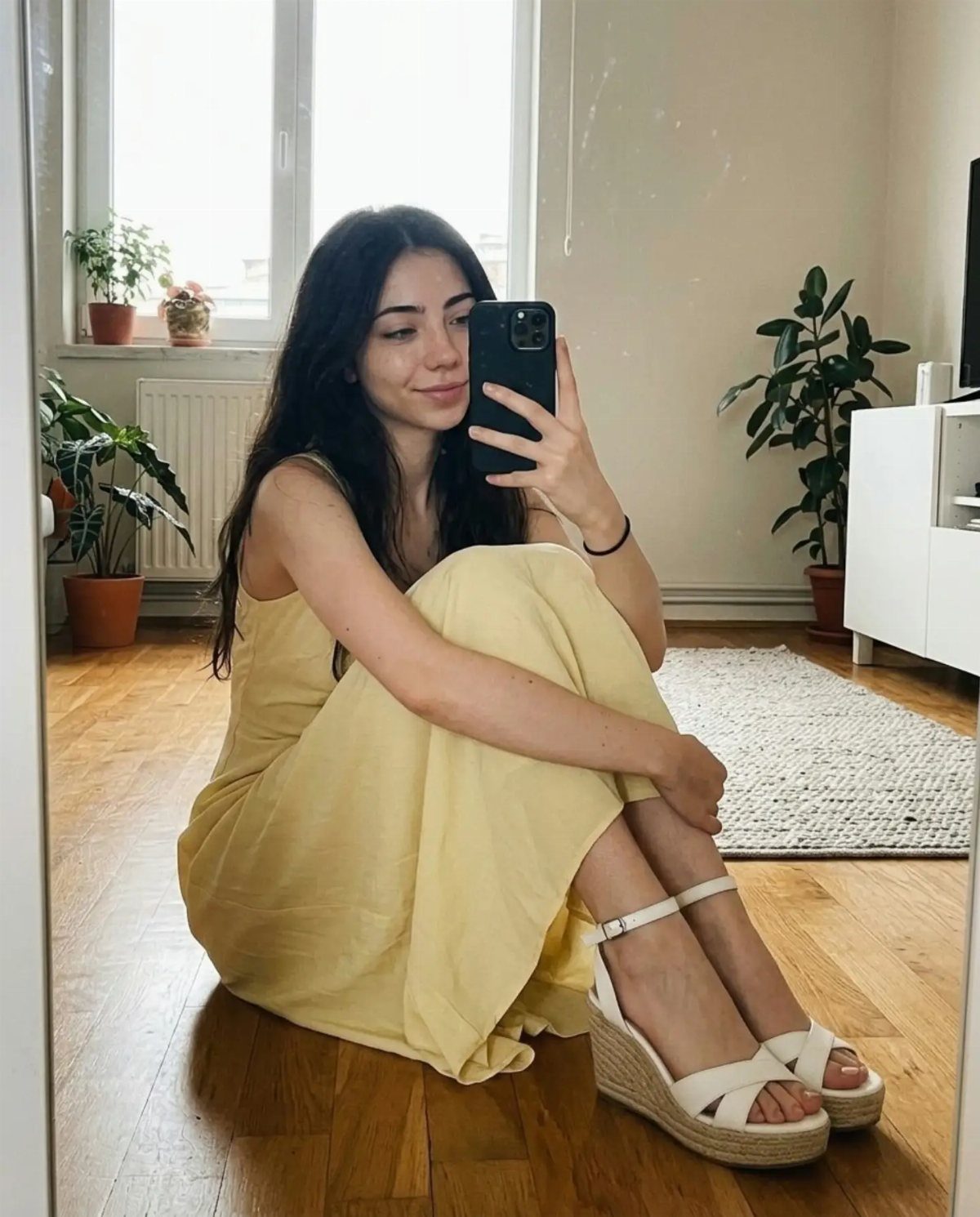 A young woman sitting on a wooden floor wearing a yellow sundress and white wedge sandals.