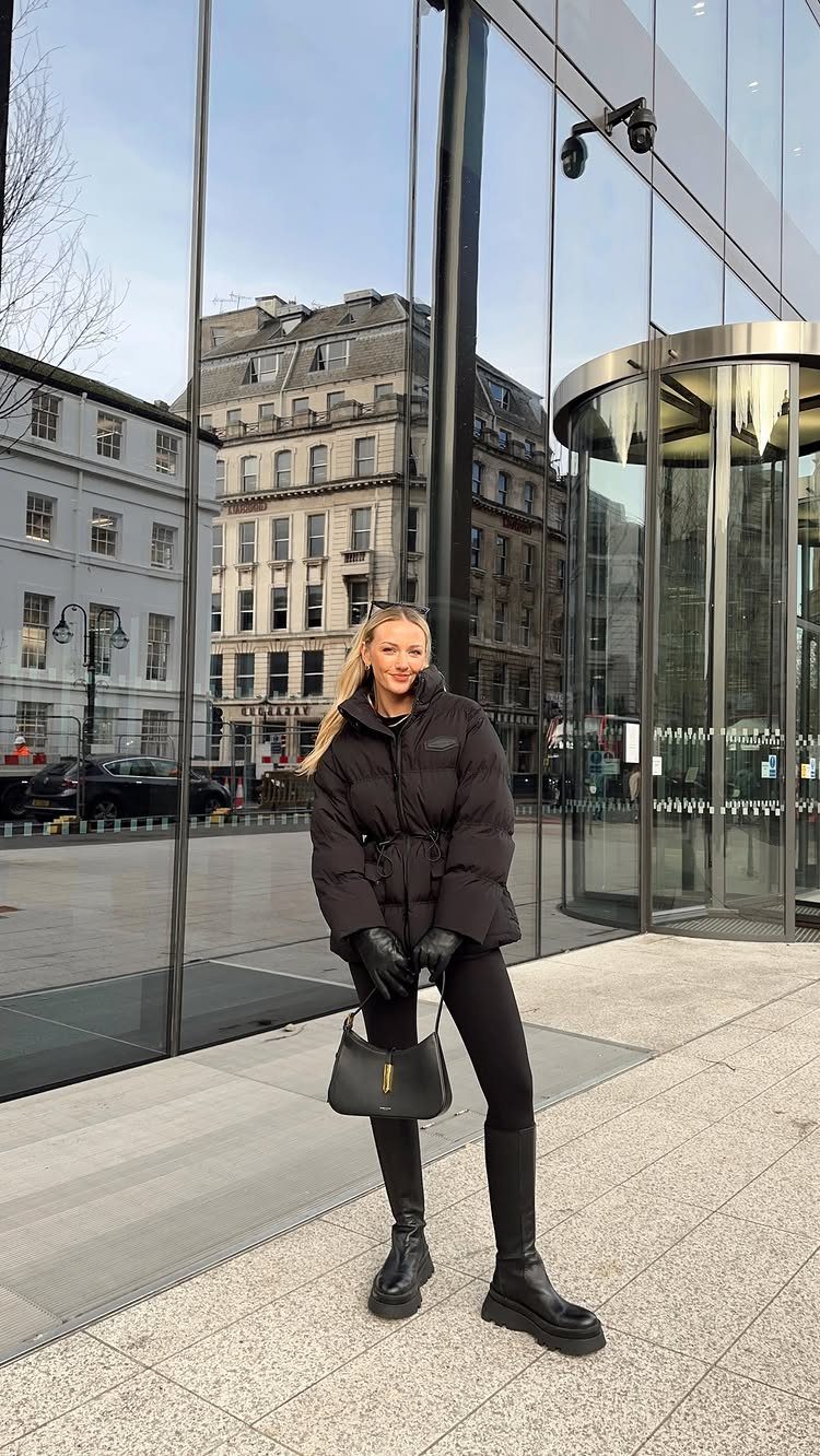 A woman in a black puffer jacket, leggings, and chunky boots standing by a glass building.