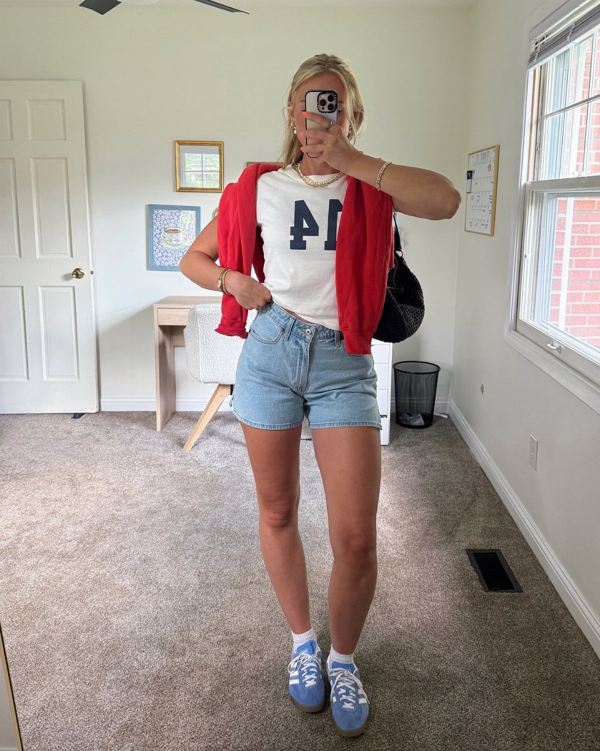 Woman in a white graphic t-shirt, red sweater, denim shorts, and blue sneakers.