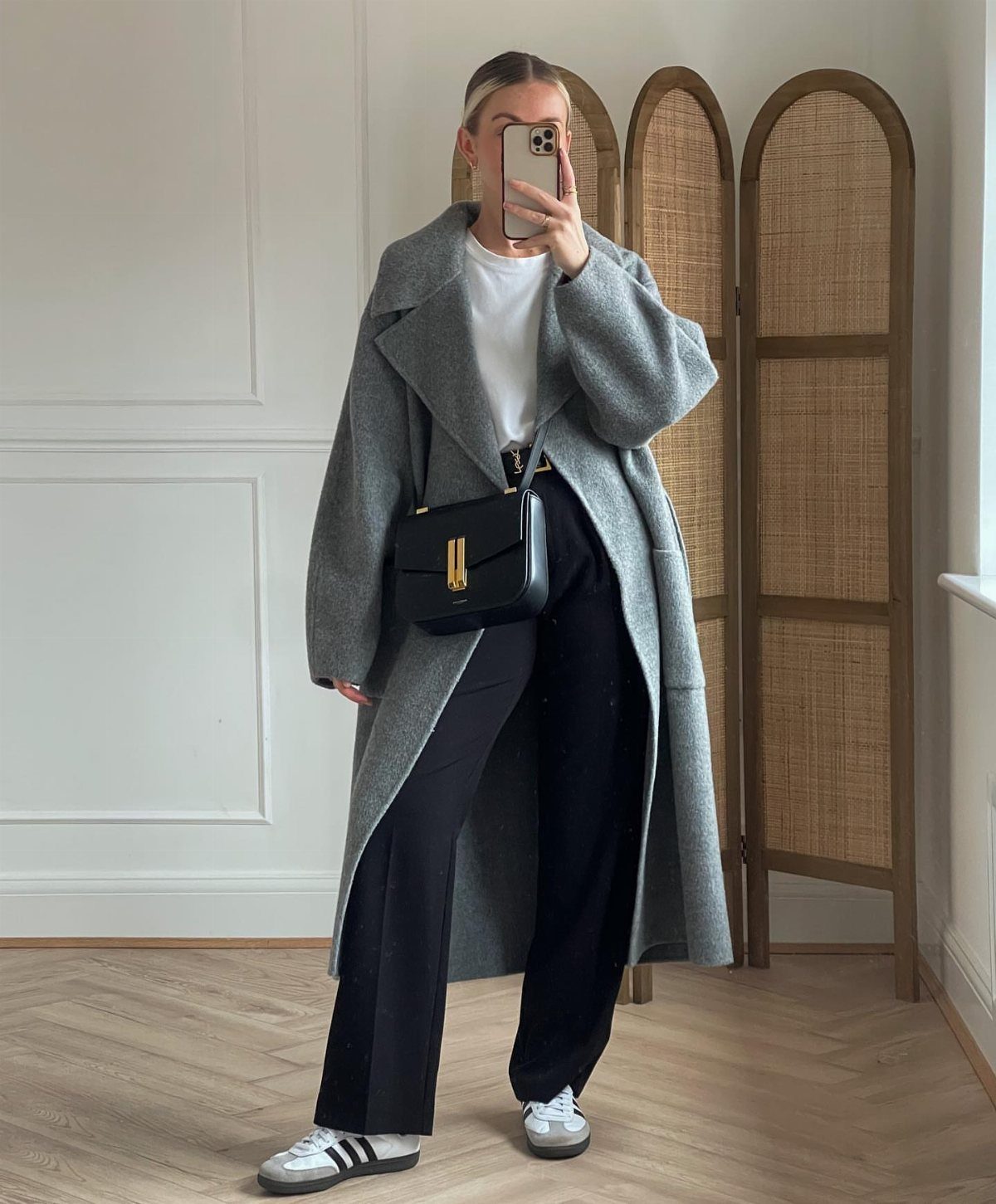 Woman in a long grey coat, white t-shirt, black trousers, and sneakers taking a selfie.