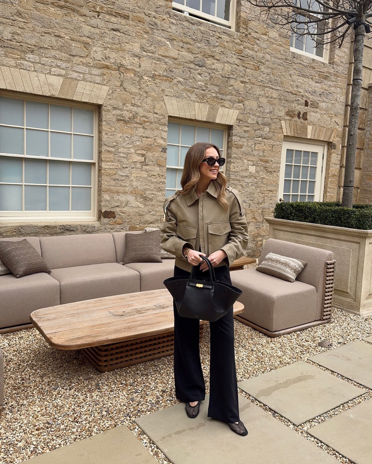 Woman wearing a tan jacket, black wide-leg trousers, and black ballet flats standing in a courtyard.