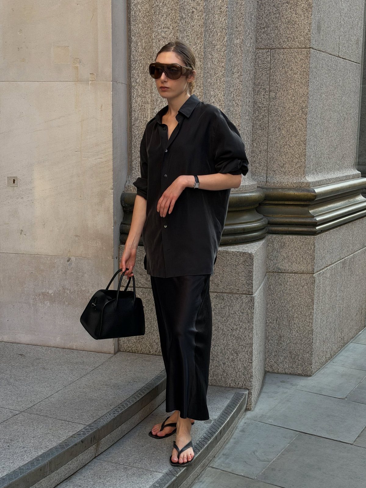 Woman wearing a black oversized shirt, satin skirt, and flip-flops standing by stone columns.