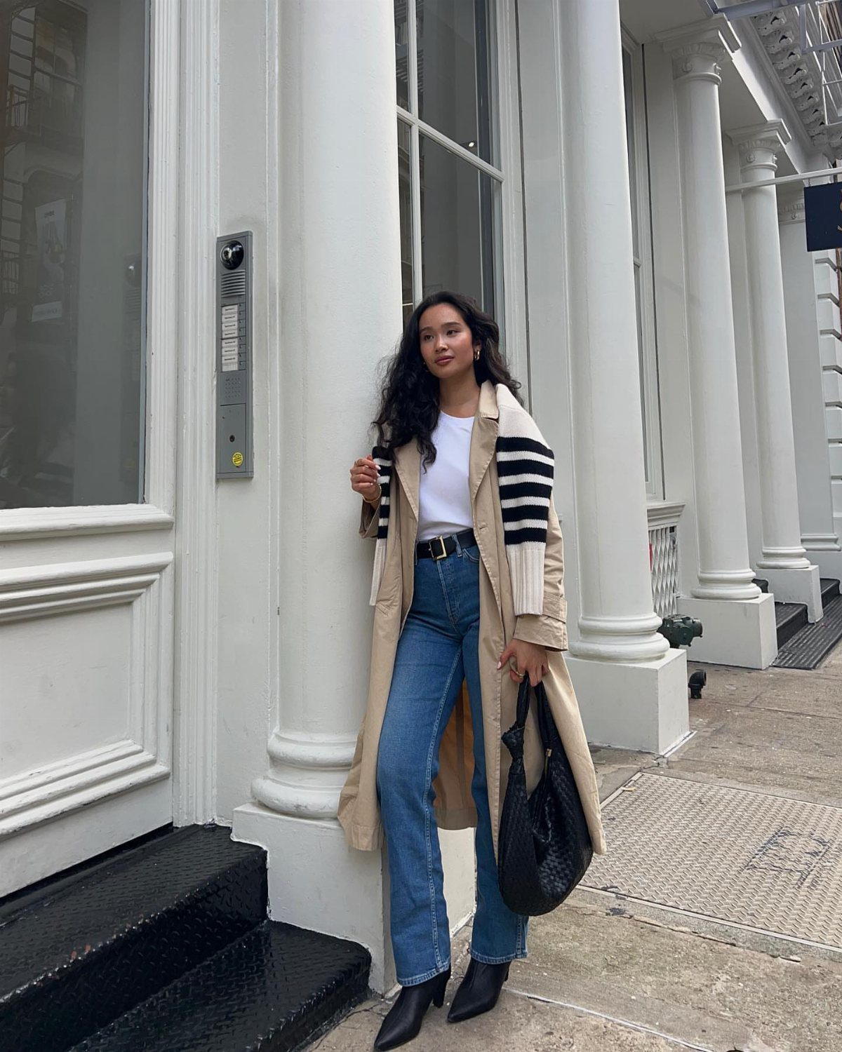 Woman wearing a long tan trench coat, white top, and blue jeans standing by white columns.
