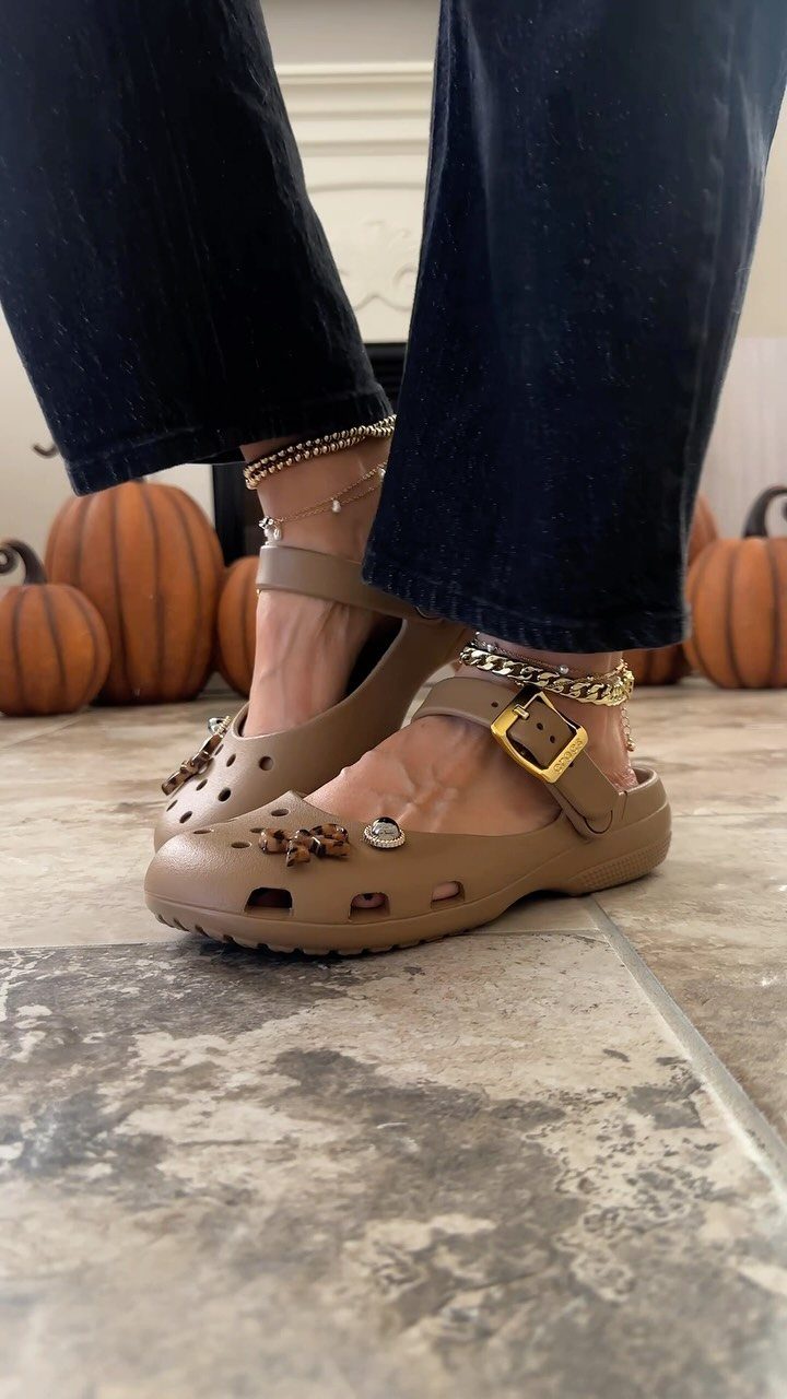 Tan clog-style sandals with gold charms and ankle bracelets worn with dark denim jeans.