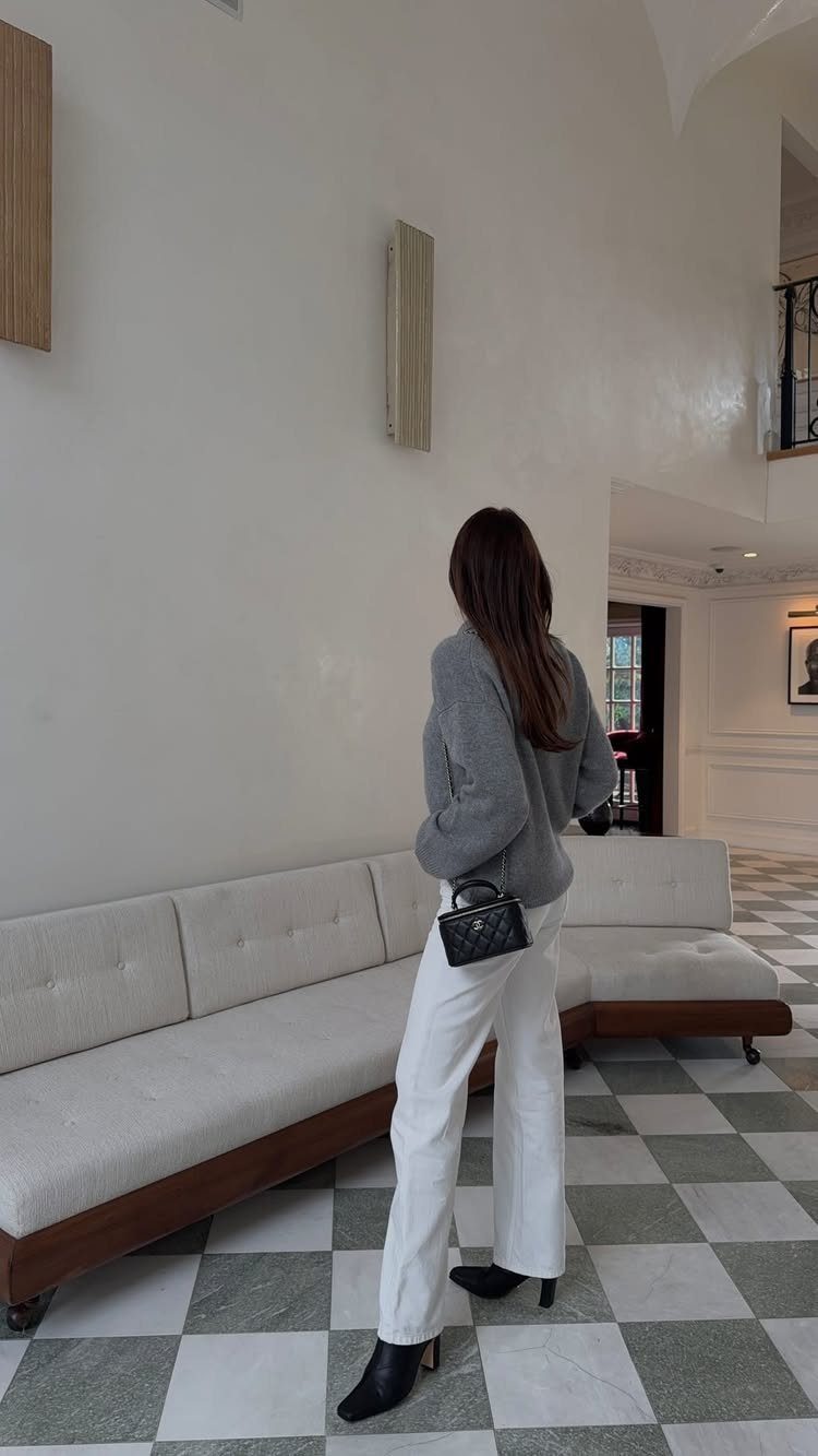 Woman in a grey sweater and white wide-leg trousers standing in a bright room.