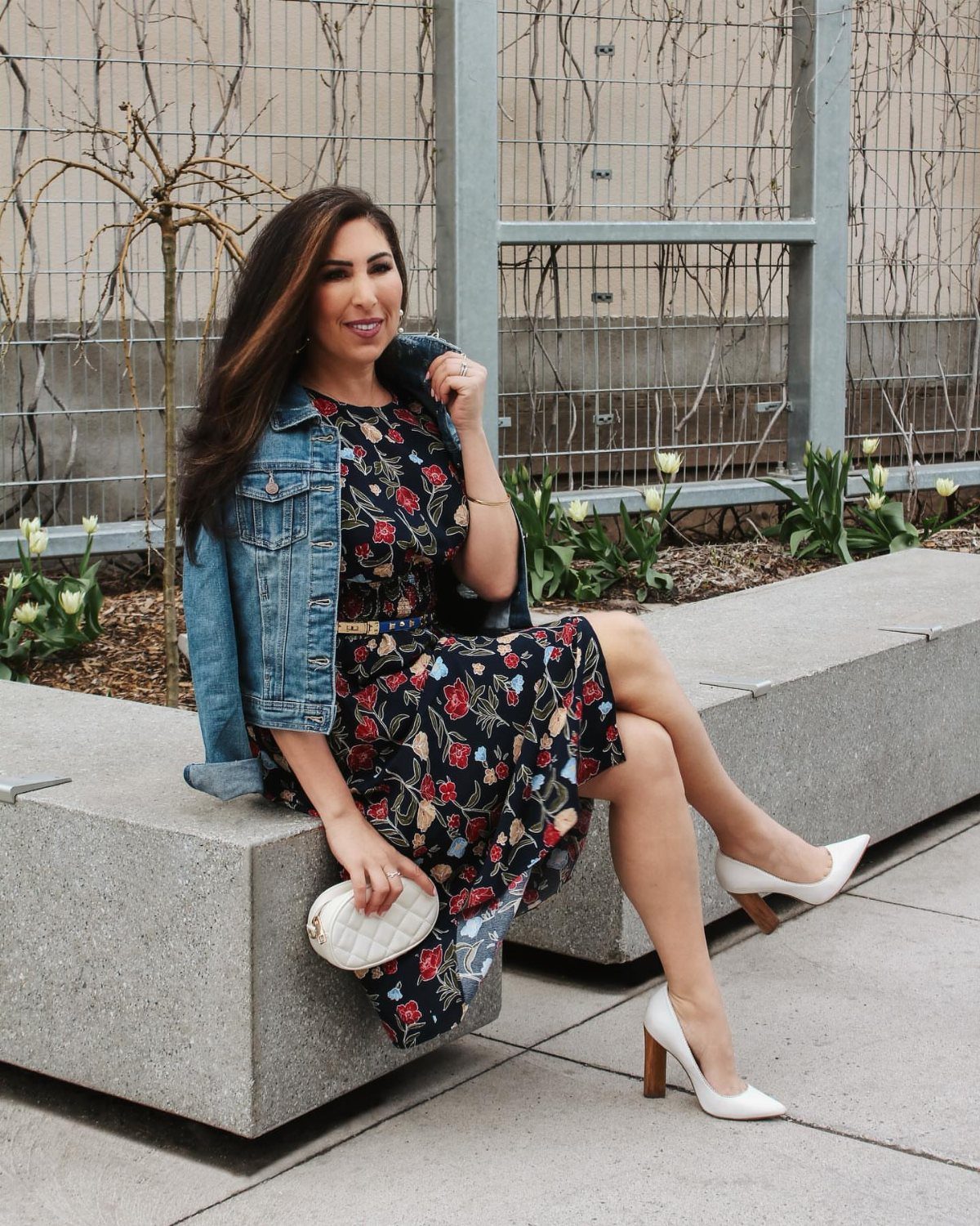 Woman sitting on a concrete bench wearing a floral midi dress and denim jacket.