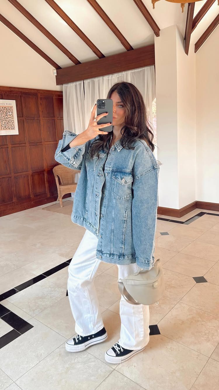 Woman in denim jacket and white trousers taking a mirror selfie indoors.
