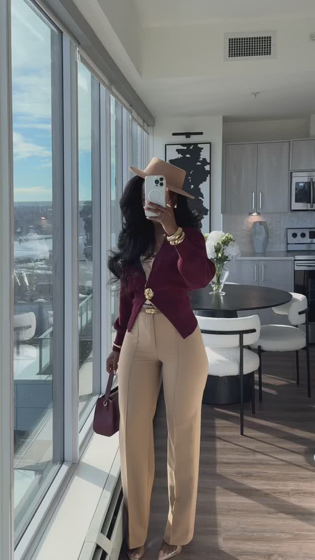 Woman in maroon cardigan and beige trousers posing for a mirror selfie in a bright apartment.