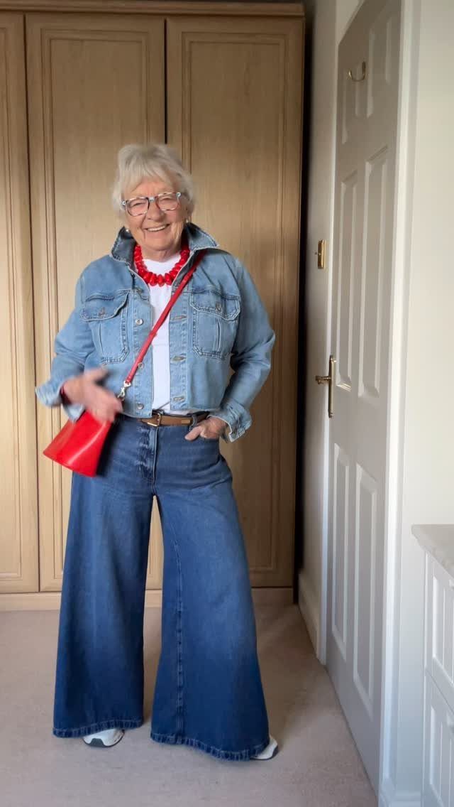 Smiling older woman wearing a denim jacket, wide-leg jeans, and a red crossbody bag.